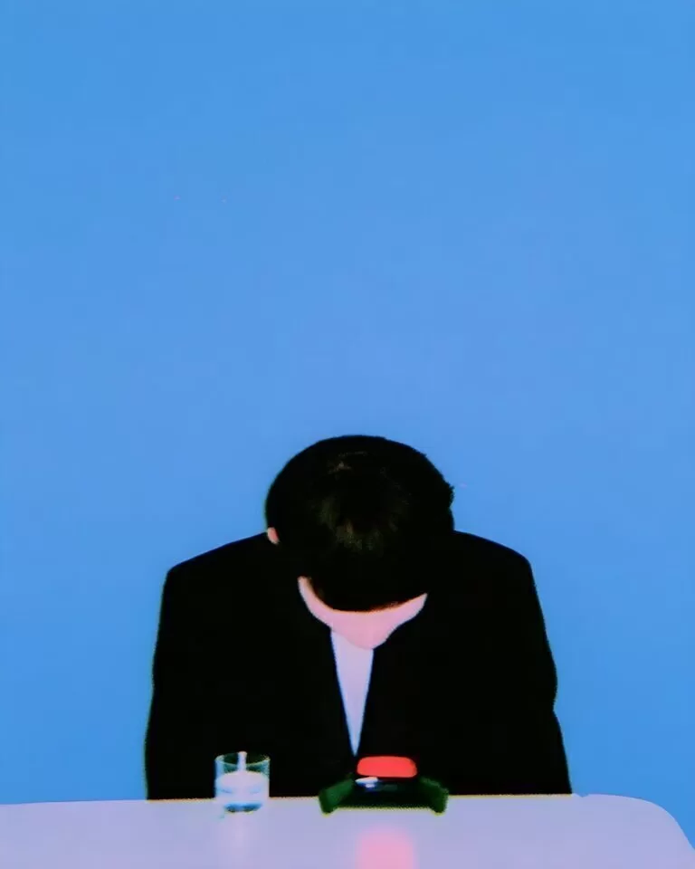 A bowed figure in a black suit sits at a white table against a vivid blue background, with a small glass and red object in front.