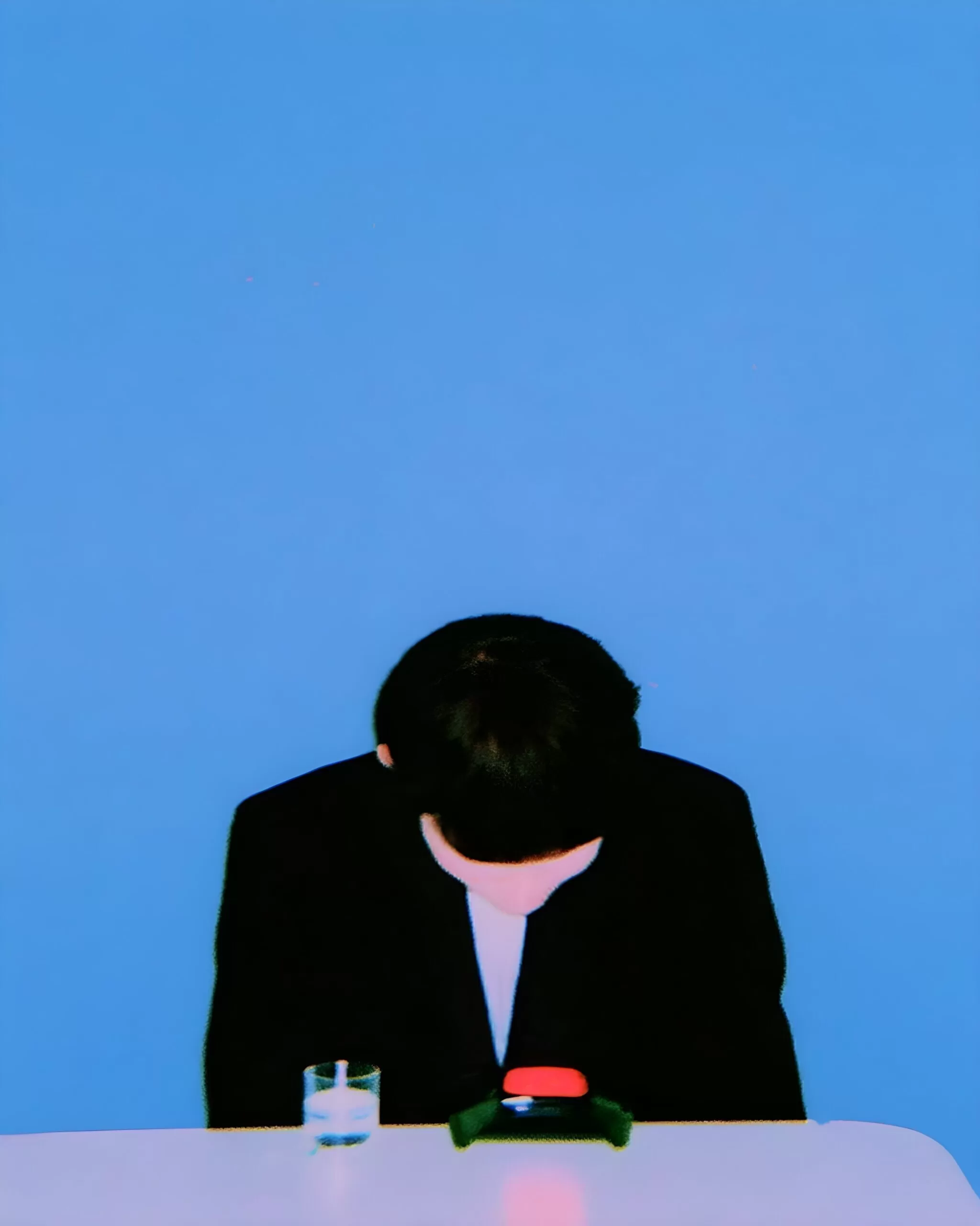 A bowed figure in a black suit sits at a white table against a vivid blue background, with a small glass and red object in front.